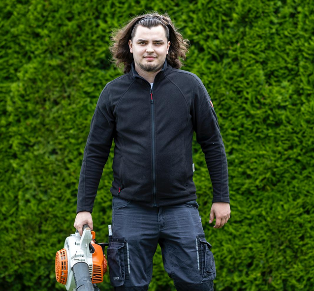 Gartenpfleger, der mit einem Laubgebläse vor einer Hecke steht.
